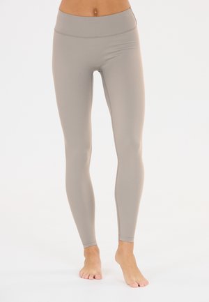 Fit kvinde iført højtaljede, lyse beige leggings i fuld længde, stående barfodet på en ensfarvet hvid baggrund.