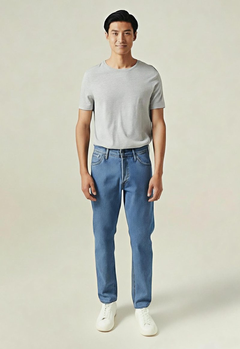 Mężczyzna stojący w jasnoszarej koszulce z krótkim rękawem, niebieskich dżinsach i białych trampkach na jednolitym beżowym tle.