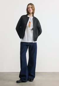 Γυναίκα που φοράει μαύρο μπουφάν τύπου bomber, λευκό μπλουζάκι με σχέδιο σκύλου, φαρδιά σκούρα τζιν παντελόνια και μαύρα παπούτσια, με λευκό απλό φόντο.