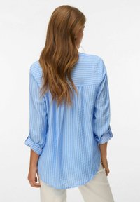 Femme aux longs cheveux bruns ondulés portant une chemise à manches longues rayée bleu clair et blanc avec les poignets retroussés et un pantalon blanc cassé, vue de dos.