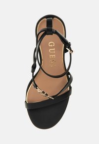 Sandale noire à talon haut avec des fines lanières, des accents en métal doré et un dessus lisse en finition mate. Semelle intérieure rembourrée couleur beige avec détail de logo.