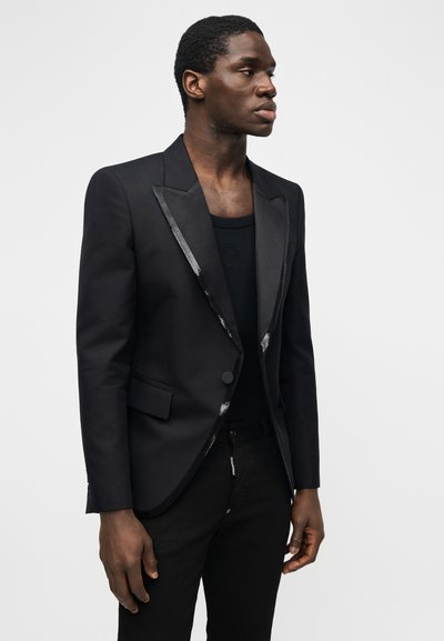 Άνδρας που φοράει εφαρμοστό μαύρο σακάκι με σατέν πέτο πάνω από μαύρο μπλουζάκι με στρογγυλή λαιμόκοψη, στέκεται μπροστά σε απλό λευκό φόντο.