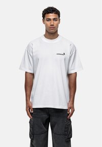 Biała, oversized koszulka z krótkim rękawem, okrągłym dekoltem i małym czarnym logo po lewej stronie. W połączeniu z ciemnymi spodniami cargo.