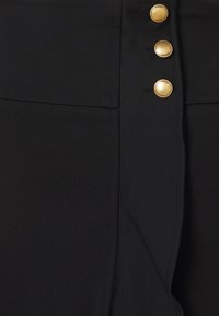 Gros plan sur un vêtement en tissu noir avec trois boutons dorés gravés du mot "PINKO" sur une couture verticale.