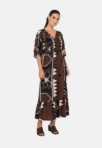 Maxikleid in Braun und Schwarz mit geometrischen und floralen Mustern, kurzen Ärmeln, V-Ausschnitt und einem geschichteten Volant-Saum. Getragen mit Blockabsatzsandalen.