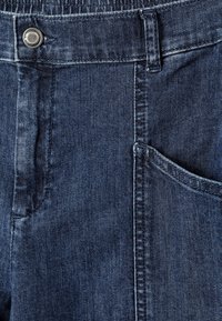 Κοντινή λήψη σκούρων μπλε τζιν με κουμπί, θηλιά για ζώνη, μπροστινή τσέπη και λεπτομερείς ραφές.