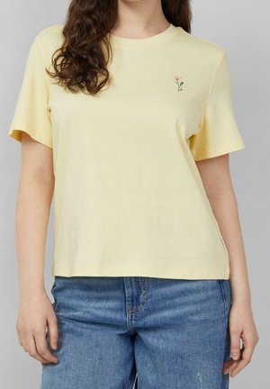 Person iført en bleg gul t-shirt med korte ærmer med en lille lyserød blomst broderet på brystet og blå jeans.