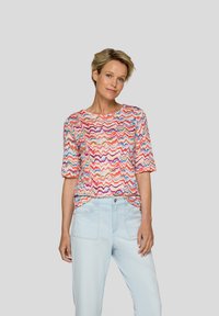 Short-sleeve T-shirt in a multicolored zigzag pattern featuring red, pink, blue, and cream hues. Fabric appears lightweight and soft.