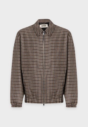 Homme portant une veste marron à carreaux, un pull beige, un pantalon en velours côtelé rose, des bottes marron foncé et un foulard bleu à motifs autour du cou, debout devant un fond blanc.