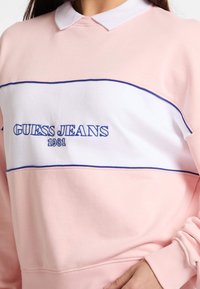 Sudadera de color rosa claro con banda blanca y bordado azul "GUESS JEANS 1981". Presenta un cuello con solapa y puños acanalados.