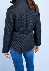 Schwarze gesteppte Jacke mit tailliertem Gürtel, hohem Kragen und langen �Ärmeln. Glattes Material mit diagonalen Nähdetails auf dem Rücken.