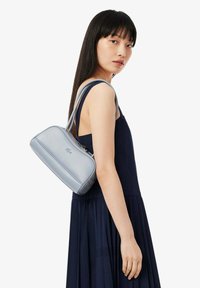 Giovane donna con lunghi capelli scuri che indossa un vestito senza maniche di colore navy e porta una borsa a spalla grigia chiara con un logo di coccodrillo.