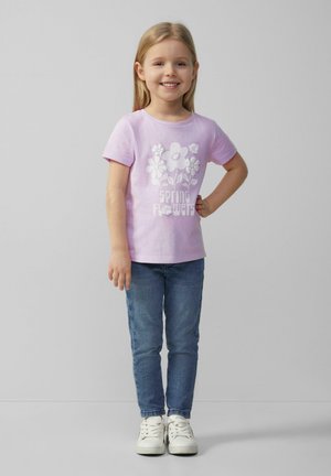 Helles lila Baumwoll-T-Shirt mit weißem Blumenmuster und dem Text "Frühling Blumen", kombiniert mit blauen Jeans und weißen Sneakers. Stehende Pose.