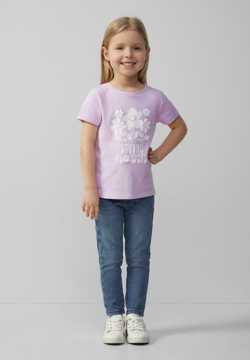 Lichtpaarse katoenen t-shirt met witte bloemenprint en de tekst "lente bloemen", gecombineerd met blauwe spijkerbroeken en witte sneakers. Staande pose.