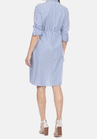 Kobieta w niebiesko-białą pionowo paskowaną sukienkę do kolan z podwiniętymi rękawami, stojąca, widoczna tyłem, w beżowych sandałach na wysokim obcasie.