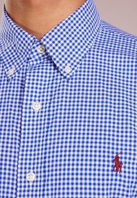 Camicia a quadri blu e bianchi con colletto button-down, che presenta un piccolo logo rosso sul petto e bottoni bianchi.