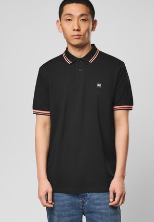 Młody mężczyzna w czarnej koszulce polo z różowymi paskami na kołnierzyku i rękawach, stojący na tle jednolitego szarego tła.