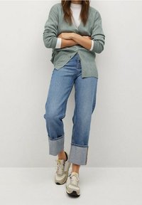 Cardigan a coste verde chiaro, camicia bianca, jeans in denim larghi con risvolti, e sneakers beige con accenti grigi.