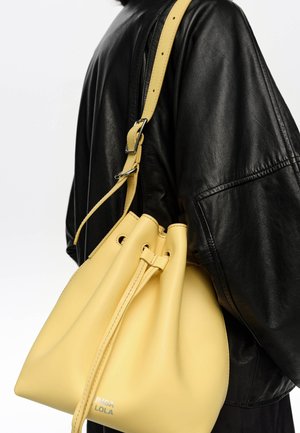 Persona con chaqueta de cuero negra llevando un gran bolso amarillo de cordón tipo cubo sobre el hombro.