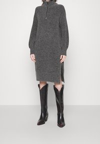 Robe tricotée grise avec un col montant et des manches longues, dotée de fentes sur les côtés. Assortie à des bottes en cuir noires de style cowboy.