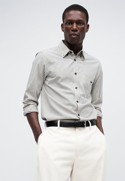 Stripete knappskjorte i lys beige og mørk brun, med en lomme, oppbrettede ermer, sammen med kremfargede bukser og et svart belte.