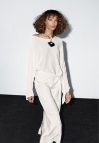 Hellgrauer, locker gestrickter Pullover mit weiten Ärmeln, kombiniert mit einem passenden langen Rock mit seitlichen Schlitzen. Eine schwarze Anhänger-Halskette vervollständigt den Look.