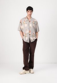 Cămașă cu mânecă scurtă, cu nasturi, de culoare gri deschis, cu modele florale și geometrice; asortată cu pantaloni largi maro și adidași într-o nuanță neutră.