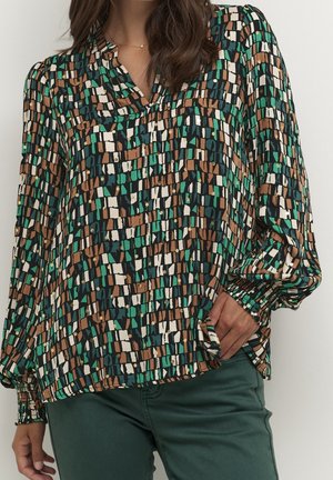Vrouw draagt een blouse met lange mouwen met abstract groen, bruin, zwart en wit geometrisch patroon en donkergroene broek, hand in de zak.