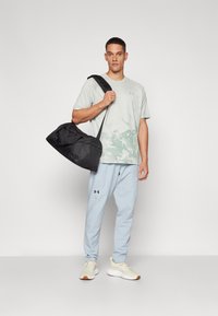 T-shirt grigio chiaro con stampa grafica verde, abbinato a pantaloni azzurri e sneakers beige. Borsa a tracolla nera. Design semplice.