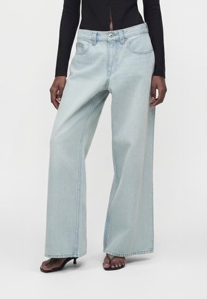 Schwarzes Langarmoberteil und hellblaue Jeans mit weitem Bein, getragen mit schwarzen offenen High Heels, Modell steht vor weißem Hintergrund.