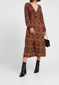 Donna che indossa un vestito a maniche lunghe con stampa tigre marrone e nera, con scollatura a V profonda, mentre tiene una borsa nera e indossa stivali neri alla caviglia.