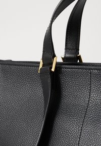 Sac en cuir noir avec une surface texturée, des accessoires en or et des poignées robustes, mettant en avant un design élégant et moderne.