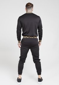 Svart träningsoverall med guldfärgad mönsterdetalj på kragen och midjebandet; figursydd design, ribbad mudd, och matchande svarta sneakers.