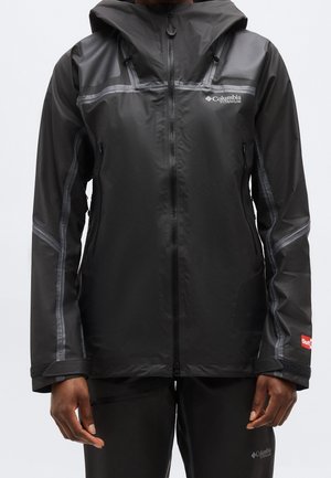 Personne portant une veste noire imperméable Columbia Titanium avec capuche et fermeture éclair, debout les bras détendus le long du corps devant un fond uni.