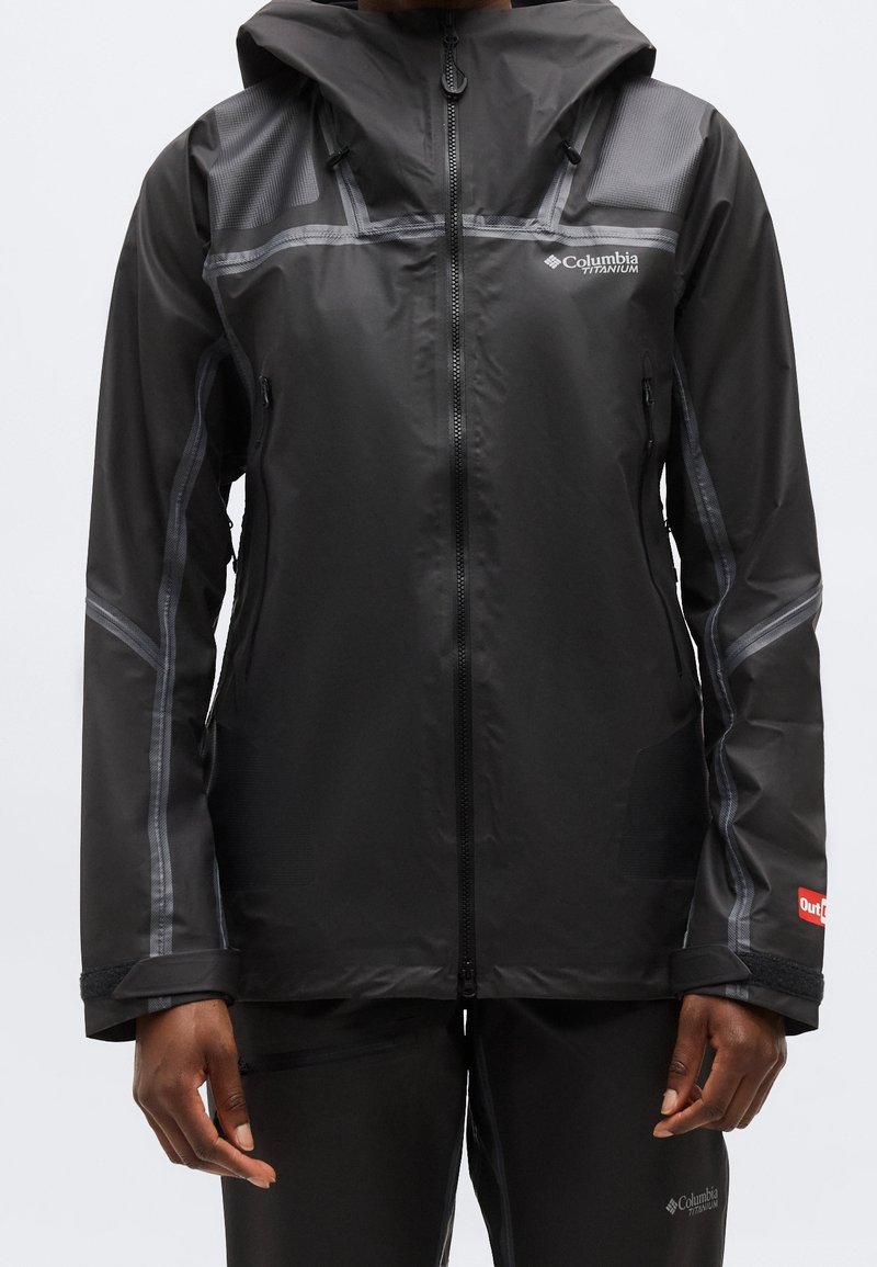 Personne portant une veste noire imperméable Columbia Titanium avec capuche et fermeture éclair, debout les bras détendus le long du corps devant un fond uni.