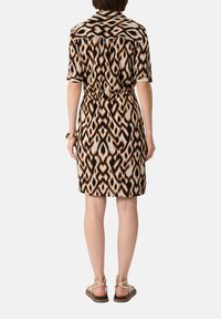 Kurzärmliges Kleid mit einem kontrastierenden geometrischen Muster in Braun, Schwarz und Creme; der Stoff ist an der Taille gerafft; kombiniert mit flachen Sandalen.