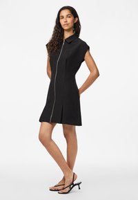 Vestido preto com gola, mangas curtas estilo manga balão, fecho frontal com zíper e detalhe de pregas. Combinado com saltos pretos com tiras.
