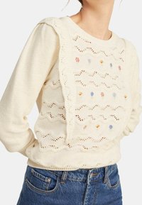 Kremowy sweter dzianinowy z falbaniastymi brzegami i haftem w kwiaty. Posiada teksturowany wzór z otwartym ściegiem. W zestawie z niebieskimi dżinsami.