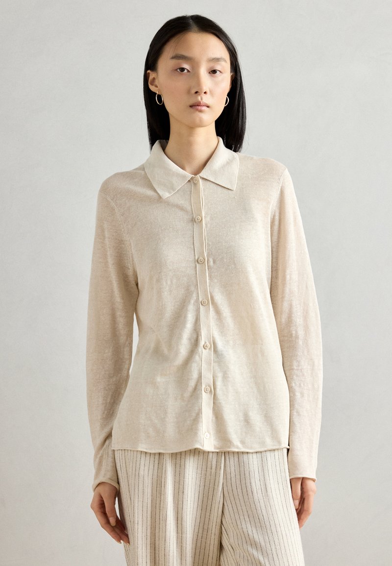 Beige skjorte med lange ermer, krage, knaplukning og subtil tekstur. Båret med lysstripede bukser. Enkel design.