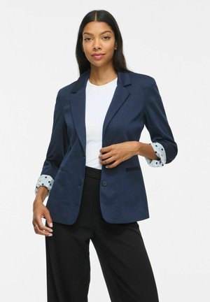 Femme portant un blazer bleu marine avec un détail de manche à pois, un top blanc et un pantalon noir, posant avec une main ajustant le blazer.