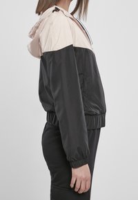 Lättvindjacka i svart och blekrosa, med huva, elastiska muddar och dragkedja fram. Materialet verkar slätt och glänsande.