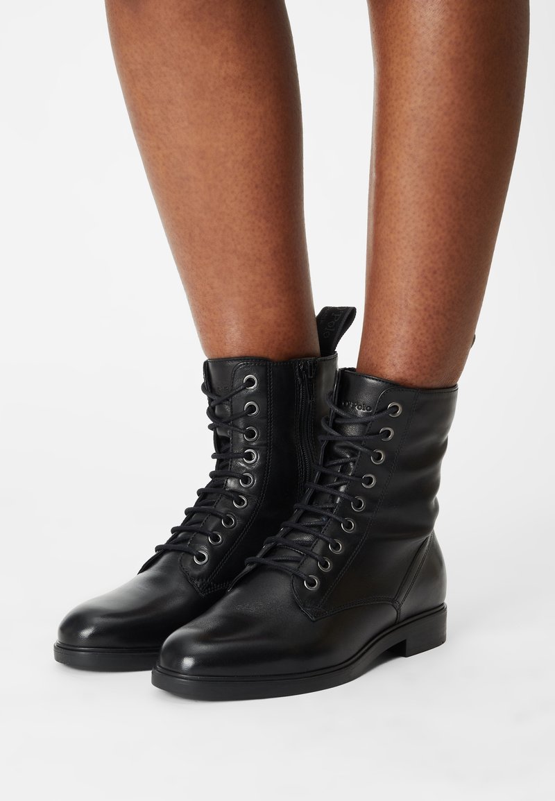 Bottes noires en cuir à lacets portées sur jambes nues sur un fond blanc uni.