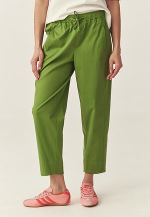 Osoba nosząca zielone spodnie ze ściągaczem i różowe trampki, stojąca na jasnym, jednolitym tle.