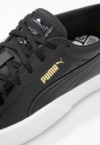 Svarta syntetiska sneakers med blank yta, med svarta snören, vit texturerad sula och guld Puma-logga på sidan.