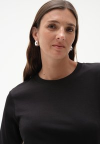 Czarna koszula z długim rękawem, okrągłym dekoltem, gładką teksturą i niewielką elastycznością. Srebrne okrągłe kolczyki odbijają światło.