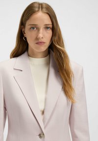Helles rosa Blazer mit strukturierten Revers und einem Knopfverschluss, kombiniert mit einem gerippten weißen Oberteil. Klare Textur und tailliertes Design.