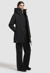 Eine Frau steht mit einem schwarzen, kapuzengefütterten Wintermantel, schwarzen Cargo-Hosen und schwarzen Schuhen vor einem einfarbigen, hellen Hintergrund.