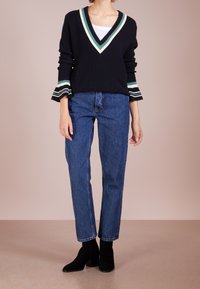 Námořnicky modrý pletený pulovr s výstřihem do V a bílými, zelenými a černými proužky; kombinovaný s modrými džíny s vysokým pasem a černými kotníkovými botami.