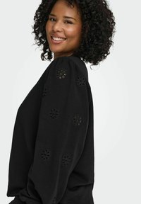 Črna dolgimi rokavi z vezenjem cvetličnih motivov na rokavih, s sproščenim krojem in mehkobo tkanine. Poudarki vključujejo okrogle izreze.