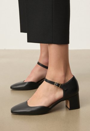 Nohy obuté v čiernych kožených topánkach s nízkym širokým opätkom, členkovými remienkami a zlatými prackami, v kombinácii s čiernymi skrátenými nohavicami.